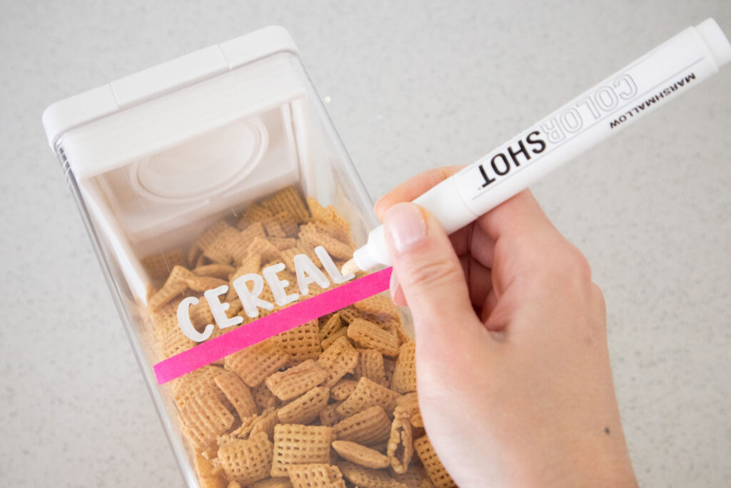 Blaire from Freshly Fuji demonstrating how to label clear food containers. There's a washi tape placed on a cereal container. She has written "cereal" using Colorshot white chalk marker.