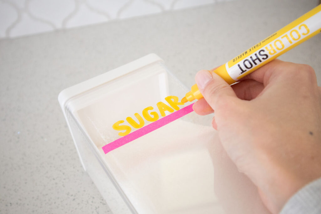 Blaire from Freshly Fuji demonstrating how to label clear food containers. There's a washi tape placed on a container of sugar. She has written "sugar" using a Colorshot yellow chalk marker.