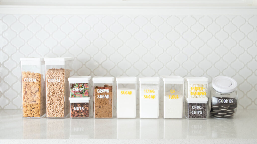 Pantry organization tip - Label clear containers using a chalk marker. Photo shows an assortment of food items in clear containers. They're all labeled with white chalk markers, except for the items that are white which are labeled with a yellow chalk marker.