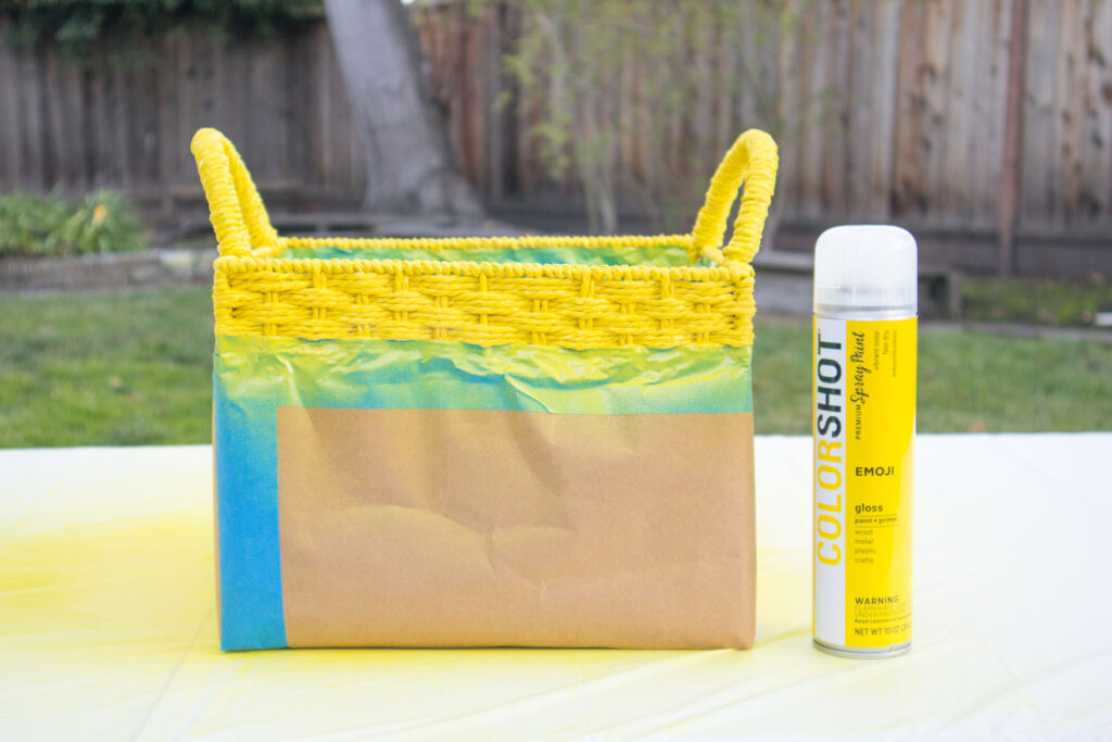 The bottom of a woven basket is taped with kraft paper and painters tape. The top section and handles are sprayed with yellow spray paint. Next to the basket is a bottle of Colorshot Emoji (yellow) spray paint.
