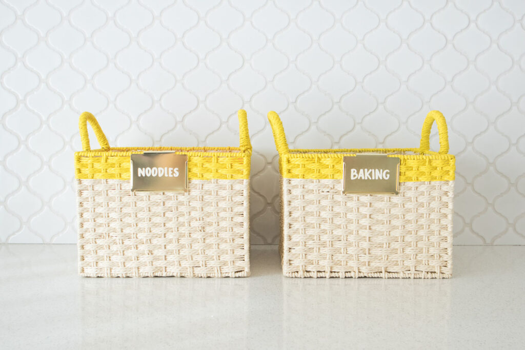 Two woven baskets with yellow stripe at the top. Each basket has a gold bin label, with "Noodles" and "Baking" written on them with Colorshot chalk marker.
