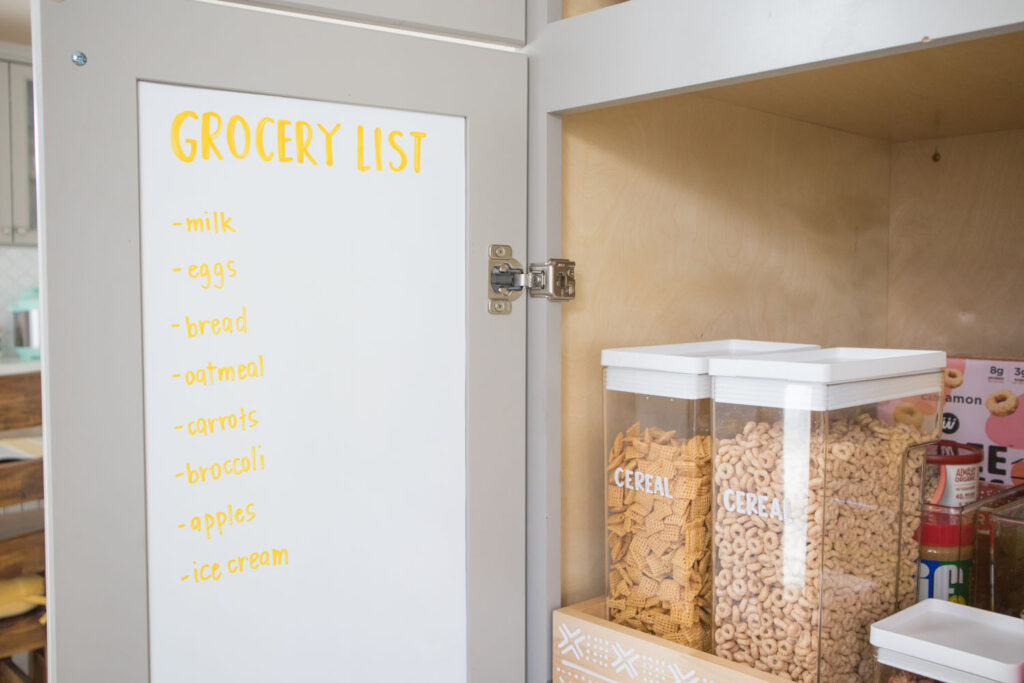 White chalkboard paper installed behind cabinet door. A grocery list is handwritten using Colorshot Emoji (yellow) chalk marker.