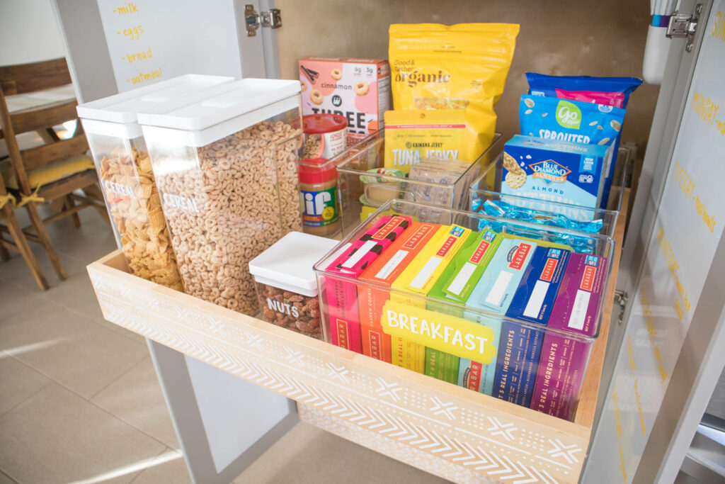 Top drawer of Blaire's pantry is pulled out slightly. It has a mudcloth print stencil on the front. Placed inside are labeled cereal containers and bins holding breakfast and snack items. The bins are painted with a section of bright yellow paint and the words are written in white chalk marker.