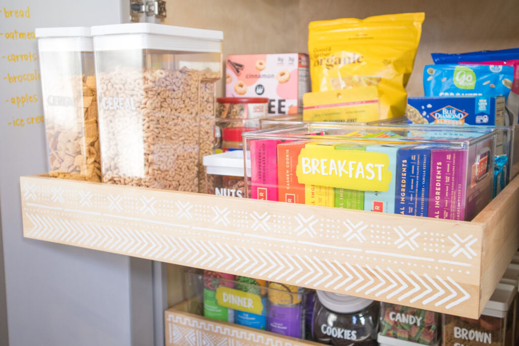 Top drawer of Blaire's pantry is pulled out slightly. It has a mudcloth print stencil on the front. Placed inside are labeled cereal containers and bins holding breakfast and snack items. The bins are painted with a section of bright yellow paint and the words are written in white chalk marker.