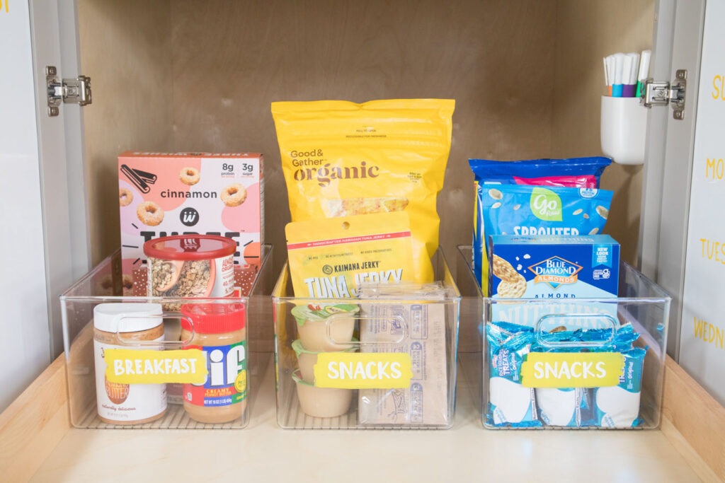 The back of the top pantry drawer is filled with clear bins. They are labeled "breakfast" and "snacks" with foods arranged in rainbow order. The labels on the bins are yellow and have the names written on them with chalk marker.