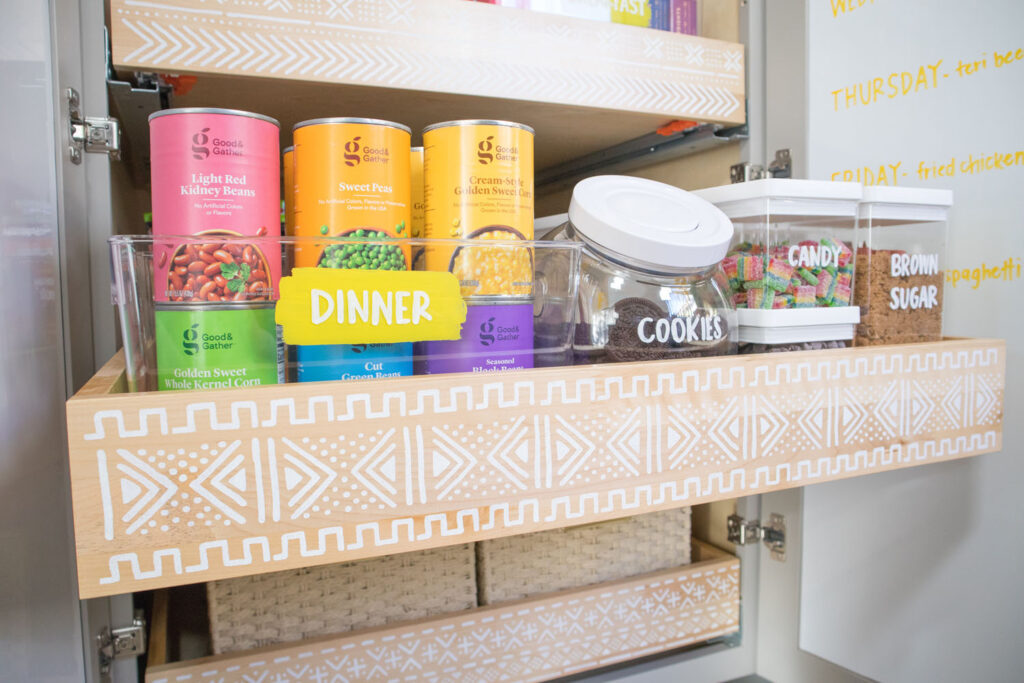 Close up of second pantry drawer. The drawer has a stenciled mudcloth design. There is a bin labeled "dinner" containing canned goods arranged in rainbow order. To the right are containers holding baking ingredients and sweets. They are also labeled with Colorshot chalk markers.