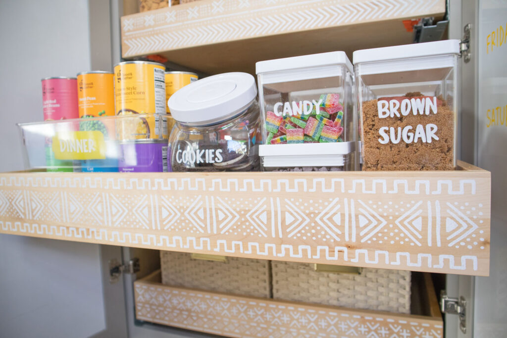 Second drawer of Blaire's pantry. The drawer is slightly pulled out and painted with a mudcloth stencil. It contains clear canisters holding cookies, candy and brown sugar. They are labeled with a white chalk marker in Blaire's handwriting. On the left is a clear bin filled with canned goods arranged in rainbow order. The bin says "dinner".