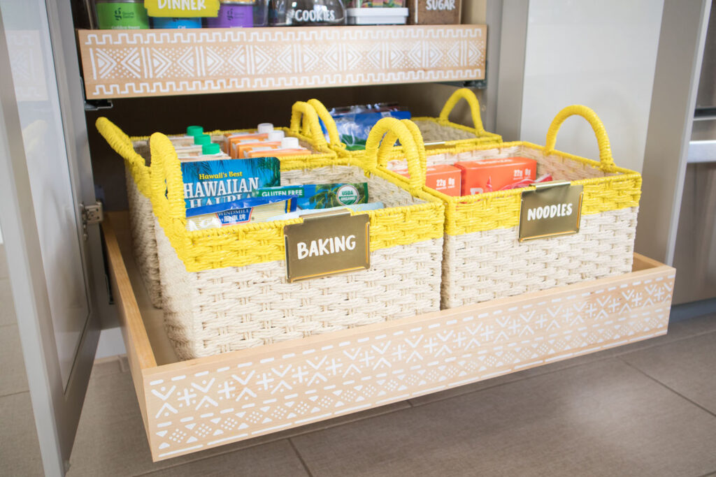 Close up of woven baskets with yellow painted stripes. Four baskets are placed on the bottom drawer of Blaire's pantry. The front baskets are labeled "Baking" and "Noodles". The drawer also has a stenciled mudcloth design on the front.