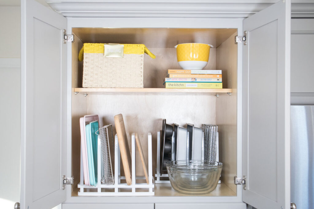 The "after" view of the top cabinet of Blaire's pantry. The top shelf holds a basket for baking pans. Next to it is a stack of yellow books and a yellow mixing bowl. The bottom portion contains white pan dividers neatly holding baking pans, cutting boards and cookie sheets.