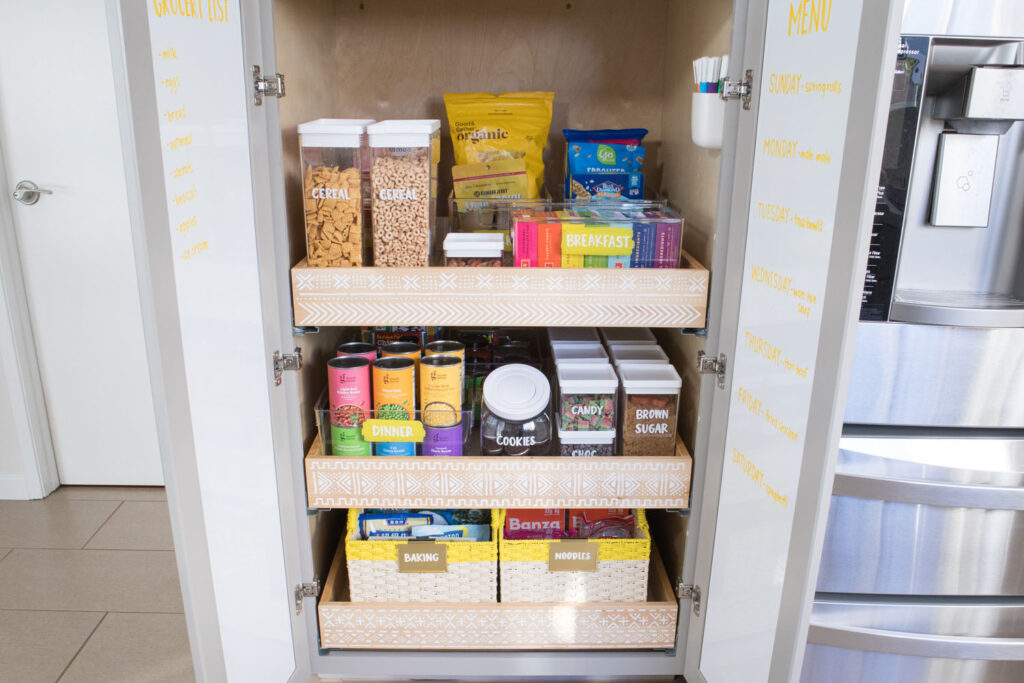 The "after" view of bottom pantry cabinet. Each drawer has a stenciled mudcloth design. The insides of each cabinet door is covered in chalkboard adhesive paper. Labeled food containers, bins and baskets are all placed neatly inside. Some of the food is arranged in rainbow order.