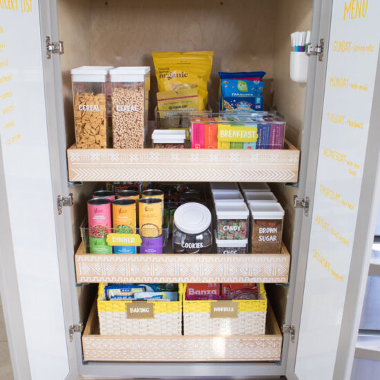 The "after" view of bottom pantry cabinet. Each drawer has a stenciled mudcloth design. The insides of each cabinet door is covered in chalkboard adhesive paper. Labeled food containers, bins and baskets are all placed neatly inside. Some of the food is arranged in rainbow order.