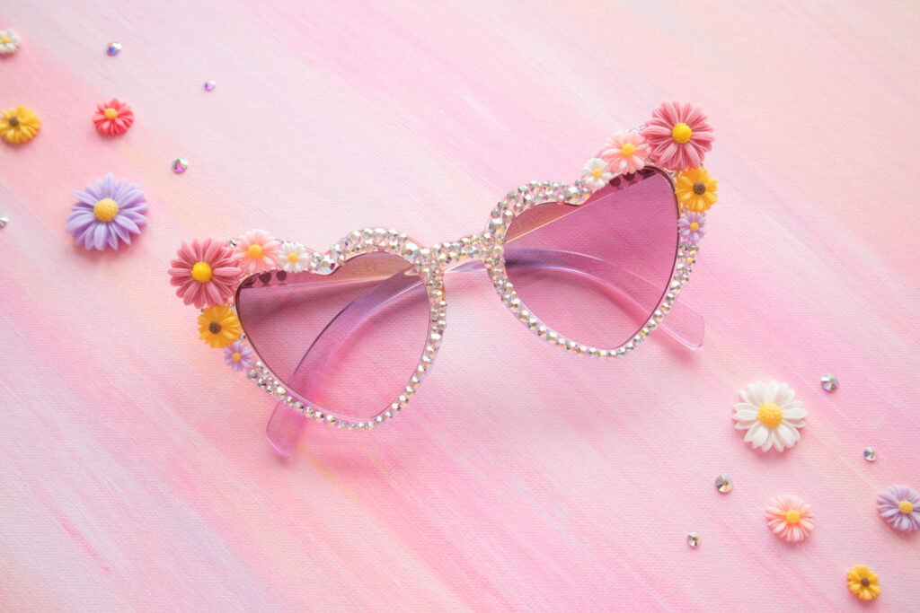 DIY rhinestone sunglasses. The sunglasses have a purple gradient and are heart shaped. They're covered in rhinestones and flower charms. The sunglasses are styled on a painted pink mat with daisy charms and rhinestones on the side.