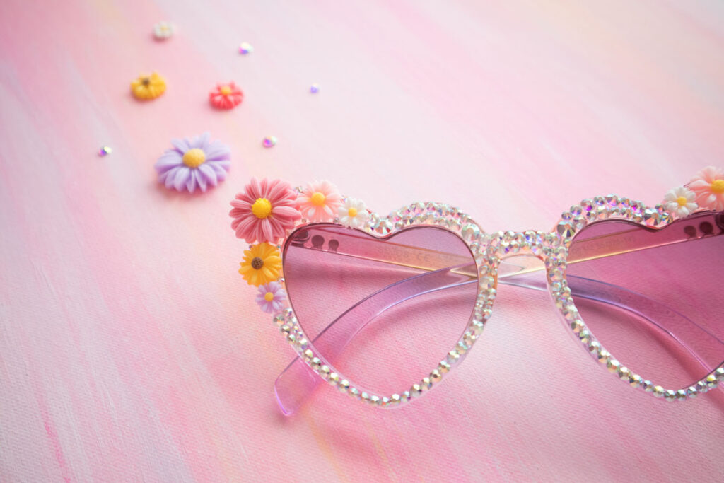 Close up of DIY rhinestone sunglasses. The sunglasses have a purple gradient and are heart shaped. They're covered in rhinestones and flower charms. The sunglasses are styled on a painted pink mat with daisy charms and rhinestones in the background.