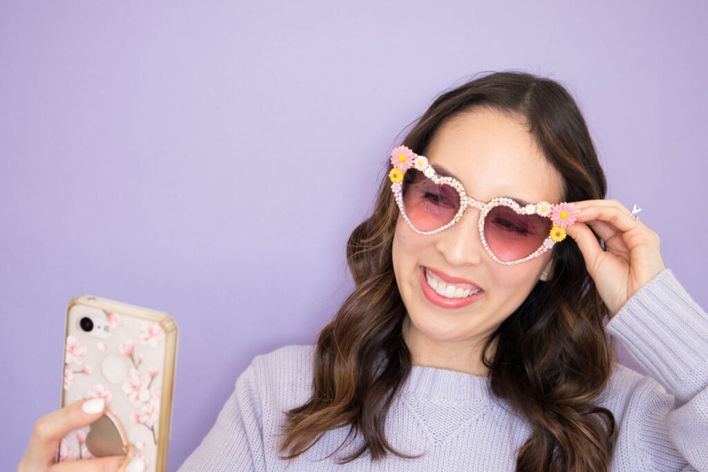 Blaire is wearing her DIY rhinestones sunglasses and is holding her phone to take a selfie. She is wearing a lavender sweater and is smiling. The background is also lavender.