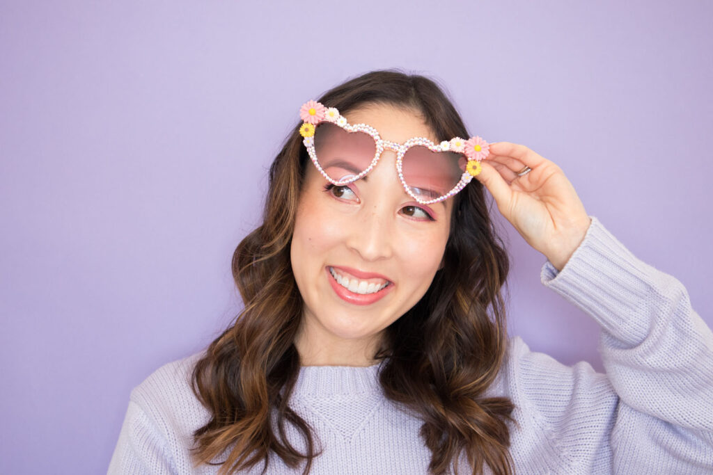 Blaire is wearing her DIY rhinestone sunglasses. She's lifting them over her eyes and smiling. She is wearing a lavender sweater and is in front of a lavender backdrop.
