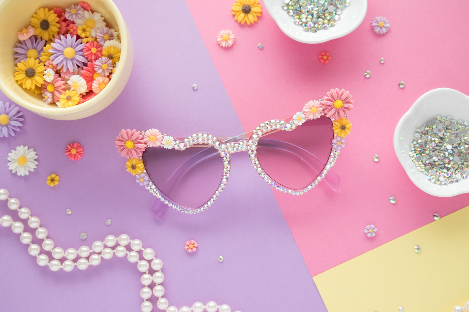 Flatlay of rhinestone sunglasses. The sunglasses are in the center. They are heart shaped and covered in rhinestones. The corners have colorful daisy charms. Surrounding the sunglasses are dishes filled with rhinestones and daisy charms. The background color blocked with lavender, pink and yellow. There are also rhinestones, flower charms and a pearl necklace loosely placed on the background.