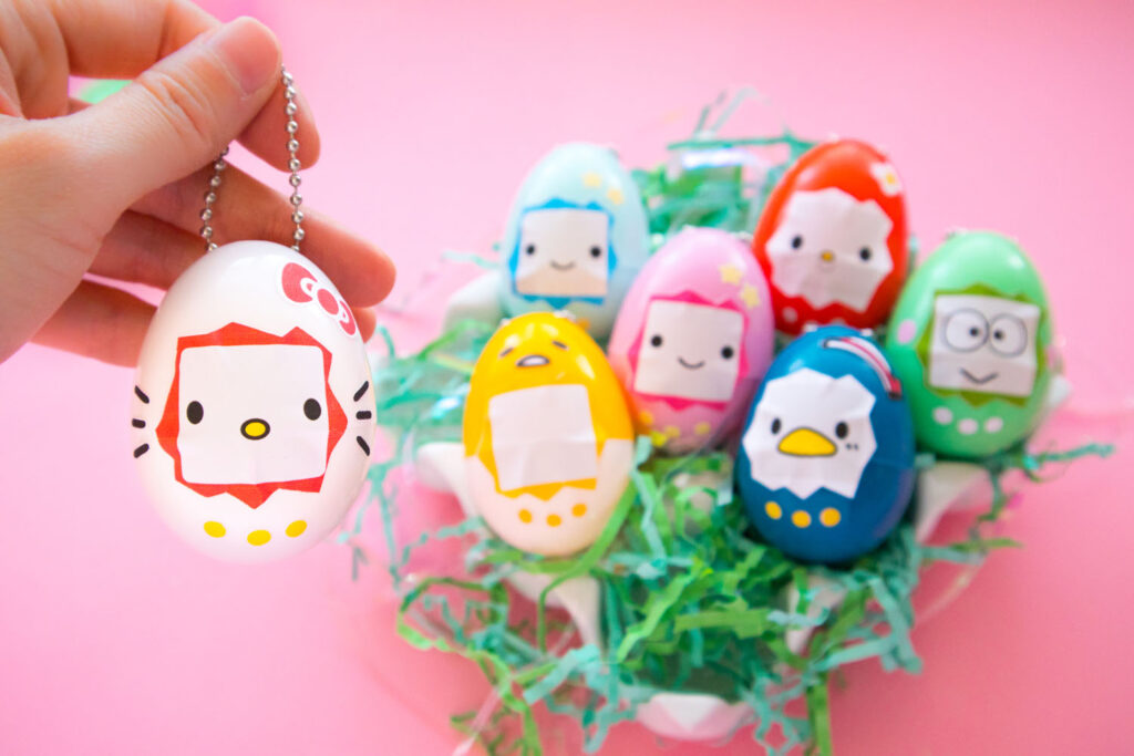 Close up shot of Hello Kitty Tamagotchi inspired egg keychain. To the right is a ceramic egg carton filled with green Easter grass and 7 plastic egg keychains make to look like the following Sanrio characters (Hello Kitty, My Melody, Keroppi, Lala, Kiki, Tuxedo Sam, Gudetama).