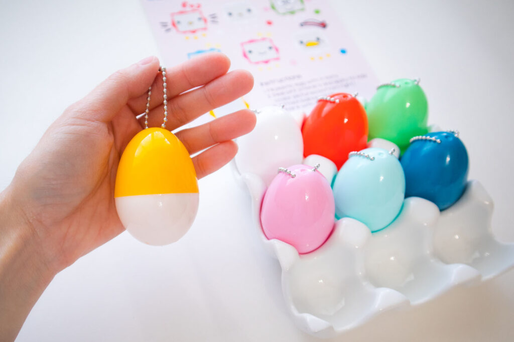 Blaire holding a half yellow and half white plastic Easter egg dangling from a ball keychain. To the right is a ceramic egg tray filled with 6 other plastic eggs with keychains attached.