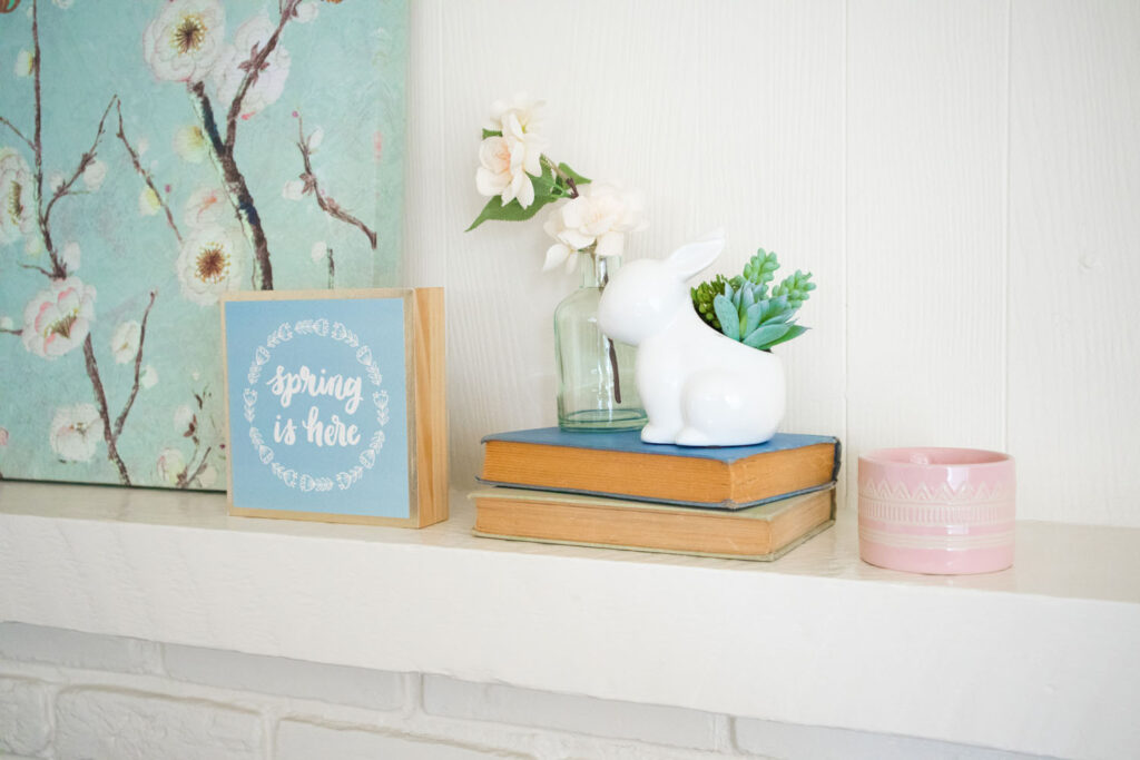 Spring living room home decor. Close up of mantel. A light blue "spring is here" sign is placed in front of the floral canvas print. To the left is a stack of two books, an aqua vase with faux flowers and a ceramic bunny planter with a succulent.