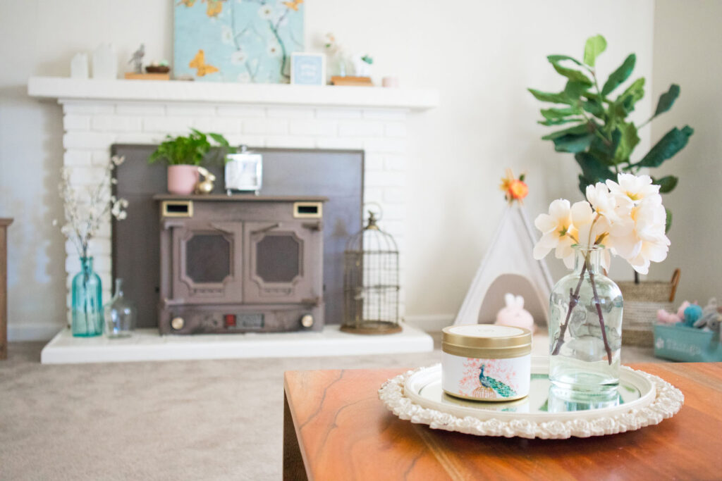 Photo of spring living room. In the foreground is a wooden coffee table with flower vase and candle propped on a mirror tray. In the background you can see the mantel decorated for spring.