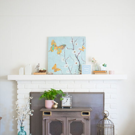 Spring living room inspiration. White brick mantel decorated for spring. Light blue canvas blossom art in center of mantel with other decorative elements.