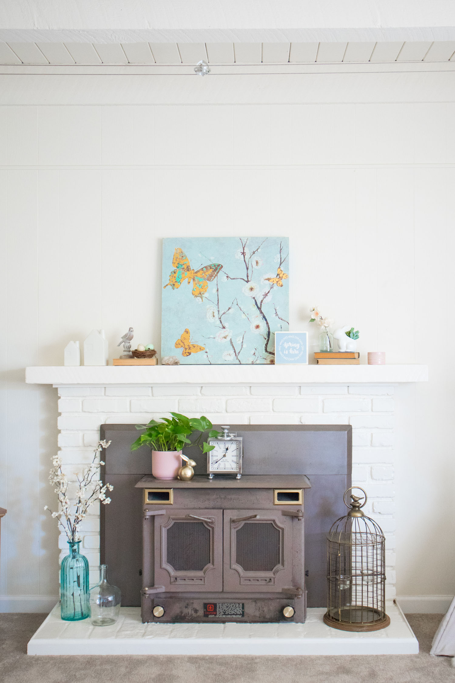 Spring living room inspiration. White brick mantel decorated for spring. Light blue canvas blossom art in center of mantel with other decorative elements.