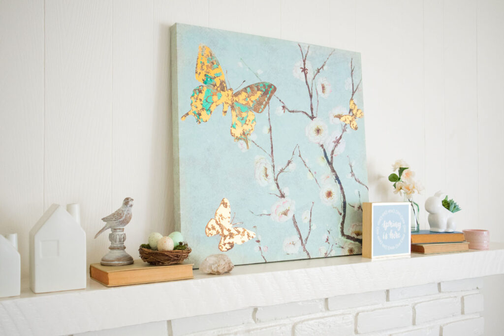 Close up of spring living room mantel. In the center is a light blue canvas with blossoms and butterflies. There are other small decorative items that match the color palette and theme.