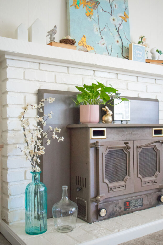 Close up of base of ivory brick fireplace. To the bottom left are glass vases. One is filled with faux blossoms. The rest of the mantel is covered in spring decor such as bunnys, birds and plants.