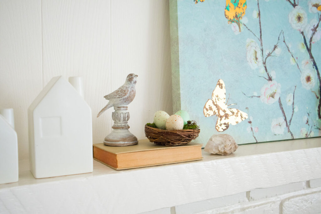 Spring living room inspiration. Close up of mantel. A wooden bird figurine and small birds nest is propped on an old book. To the right is natural stone and a blue canvas art print.