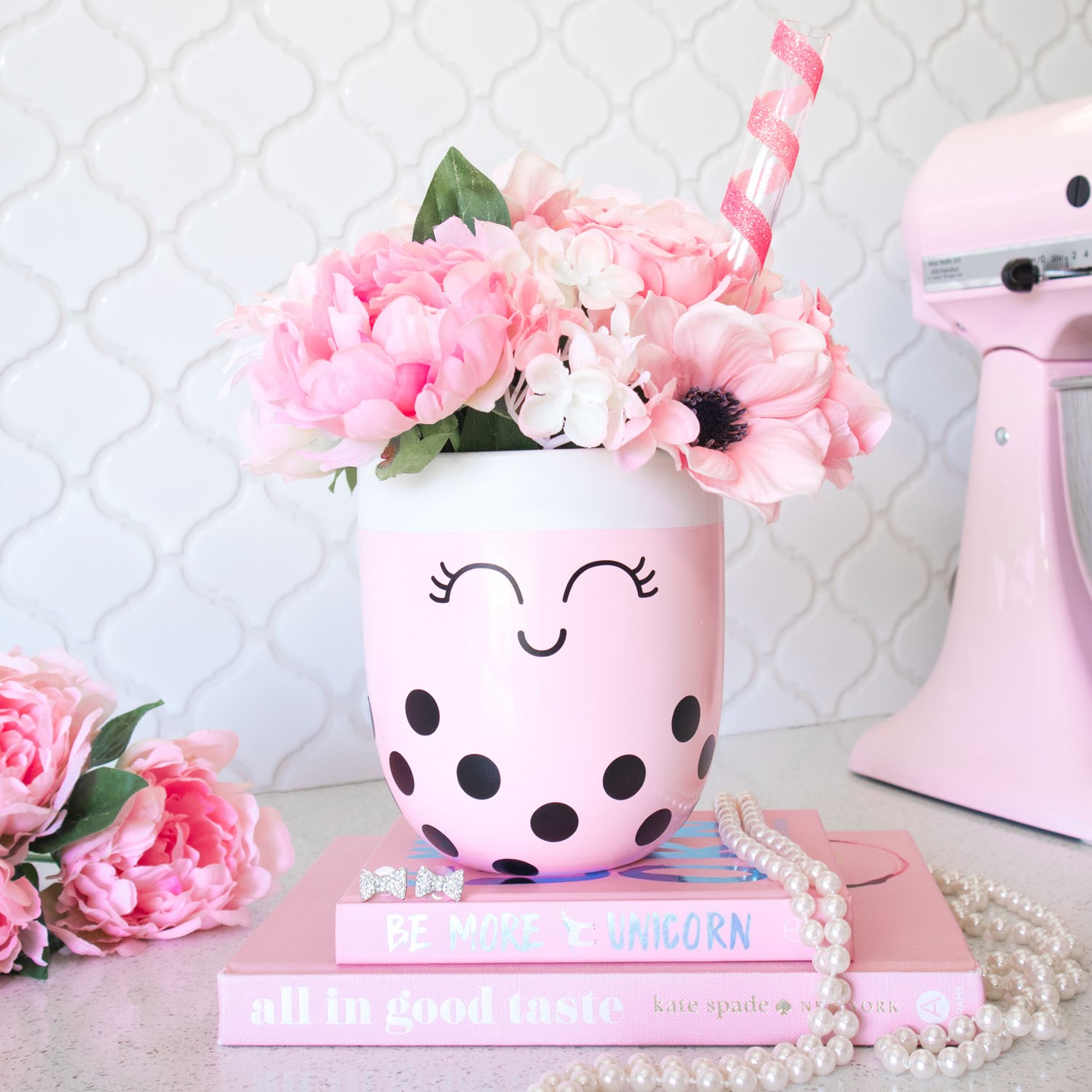 Pink boba plant pot with faux pink flowers. It's placed on a stack of pink books and styled with pearls, flowers and a pink kitchenaid mixer.
