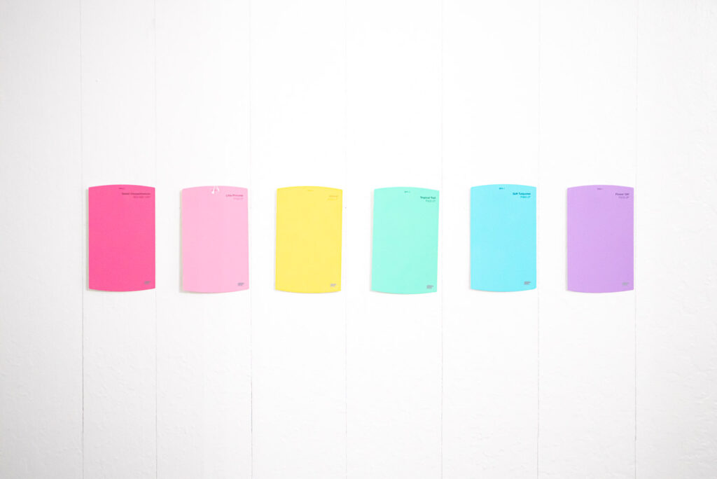 6 paint chips are arranged in rainbow order on the white wall. Penciled vertical markings are placed where stripes will be.