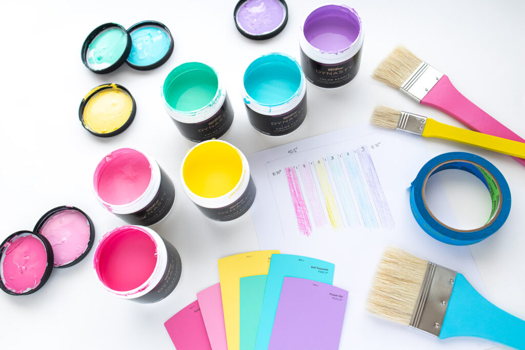 A flatlay photo of materials needed for painting rainbow stripe wall: small jars of paint (hot pink, pink, yellow, mint, turquoise, lavender), a wall sketch, paint swatches, colorful paint brushes and painter's tape.