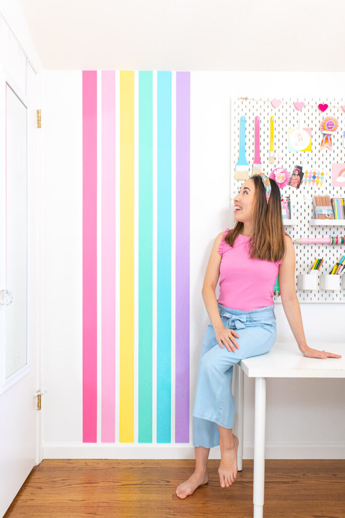 Blaire, a petite Asian American woman with brown hair is dressed in a bright pink top and light blue pants. She is sitting on her white craft room desk and looking up at her rainbow stripe wall.