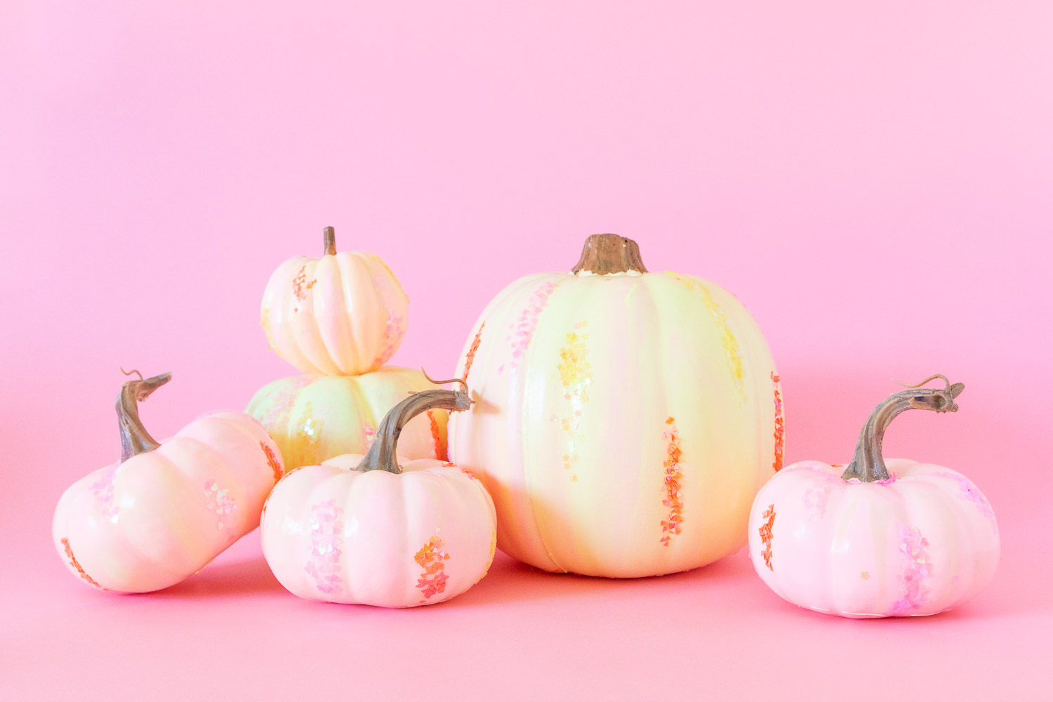 A variety of neon paint poured pumpkins with light pink, orange and yellow marbled effect. There is also pink, orange and yellow glitter attached to each pumpkin. They're displayed in a group on a pink backdrop.
