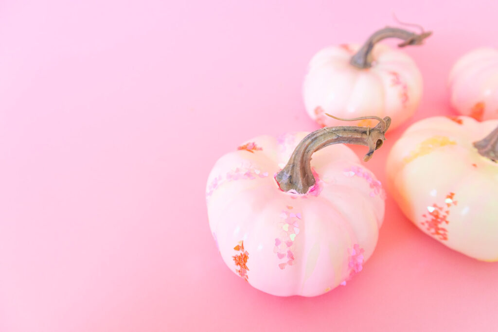 Close up of mini paint poured pumpkins. They're covered in light pink, orange and yellow swirled paint and glitter. They are placed on a pink backdrop.