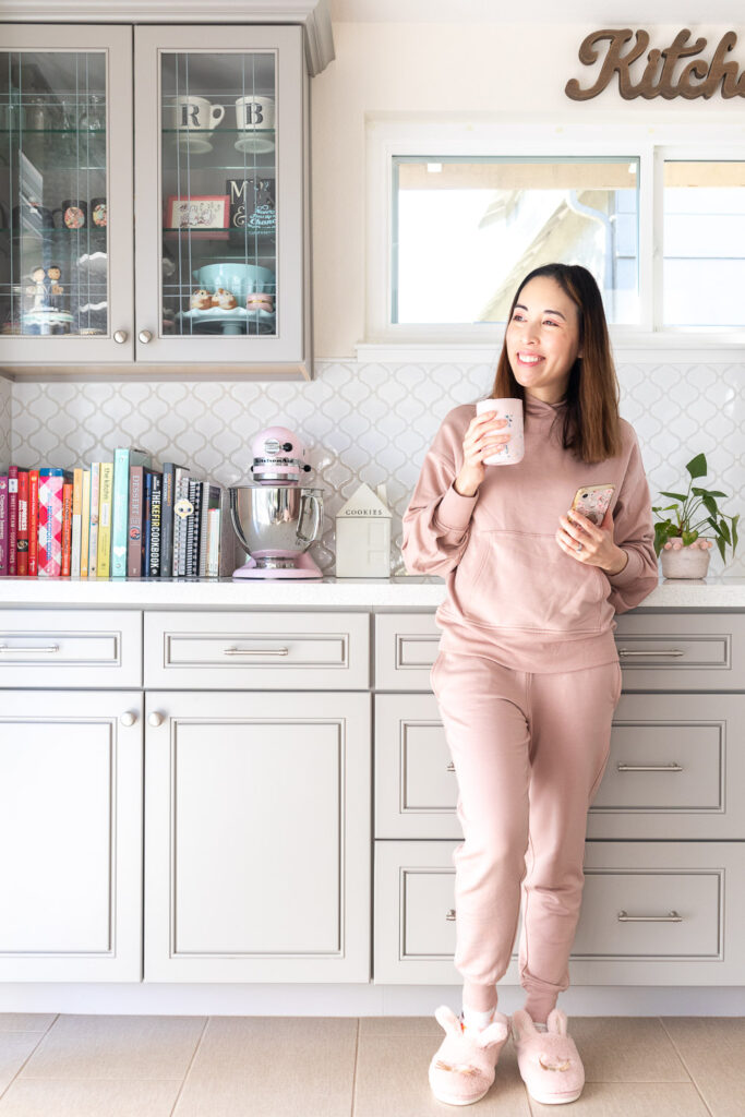 Blaire is wearing a blush hoodie and matching jogger pants. She is holding a pink travel mug and her phone in the kitchen.