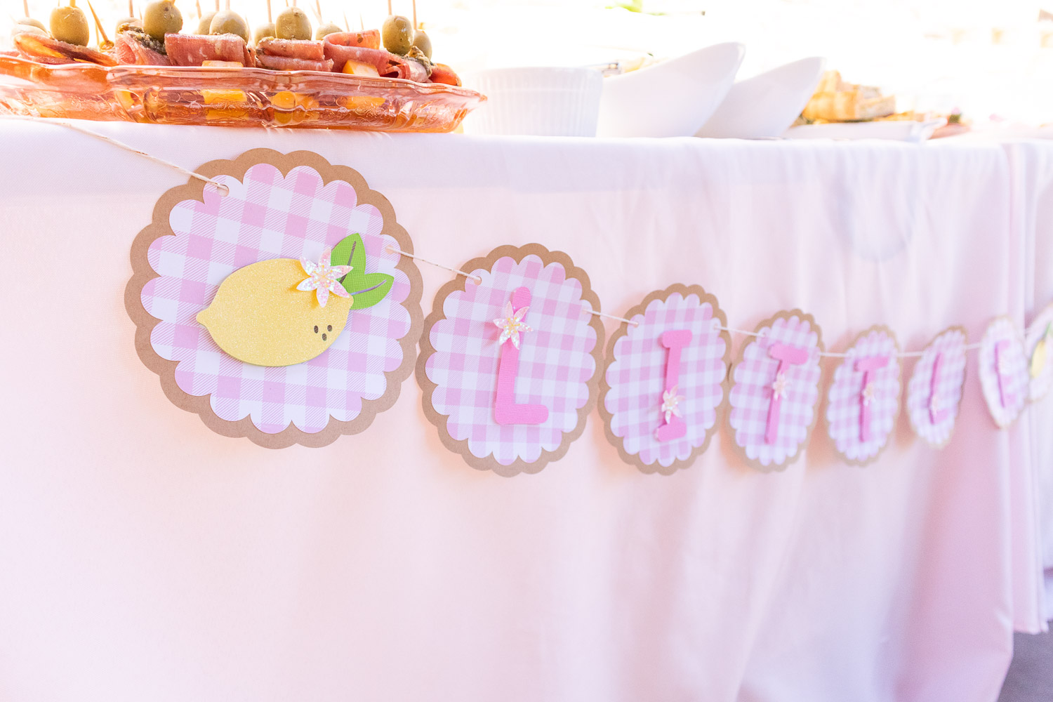 Closeup of lemon baby shower banner made with Cricut. Each pink letter is placed on scalloped gingham circles. The piece on the end has a lemon with a glittery blossom.