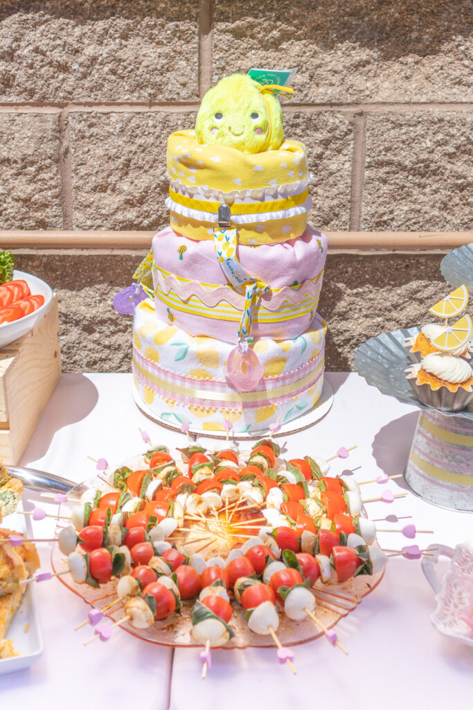 A three-tired pink, yellow and white diaper cake with a lemon plush on top. It sits in the center of the food table. 
