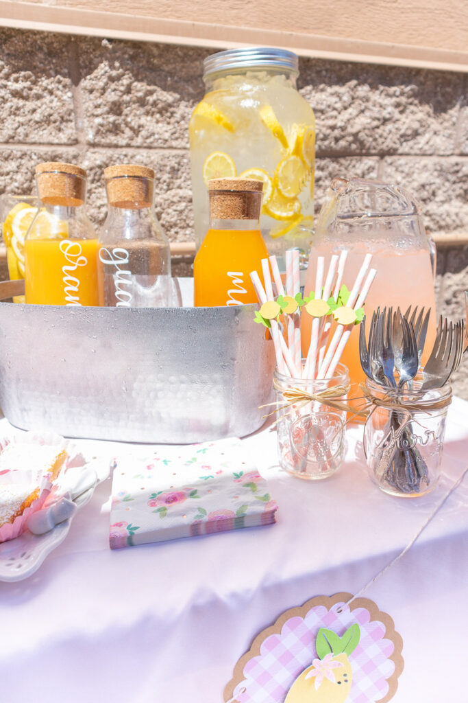 Beverage station featuring a several juices in carafes, pink lemonade in a pitcher and lemon water in a large dispenser. In front of these are handmade lemon paper straws.