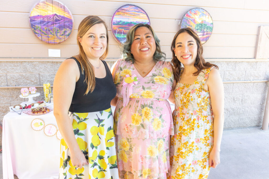 Jenna from (Heart and Arrow Weddings) and Blaire (from Freshly Fuji) with guest of honor, Joie. They are all wearing printed summer dresses and smiling. Joie's dress is pink with yellow flowers. 