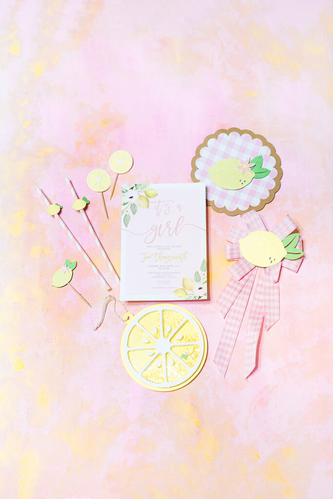 Baby girl shower invitation is light pink with yellow watercolor lemons. It is styled on a hand painted pink and yellow backdrop with other handcrafted elements.