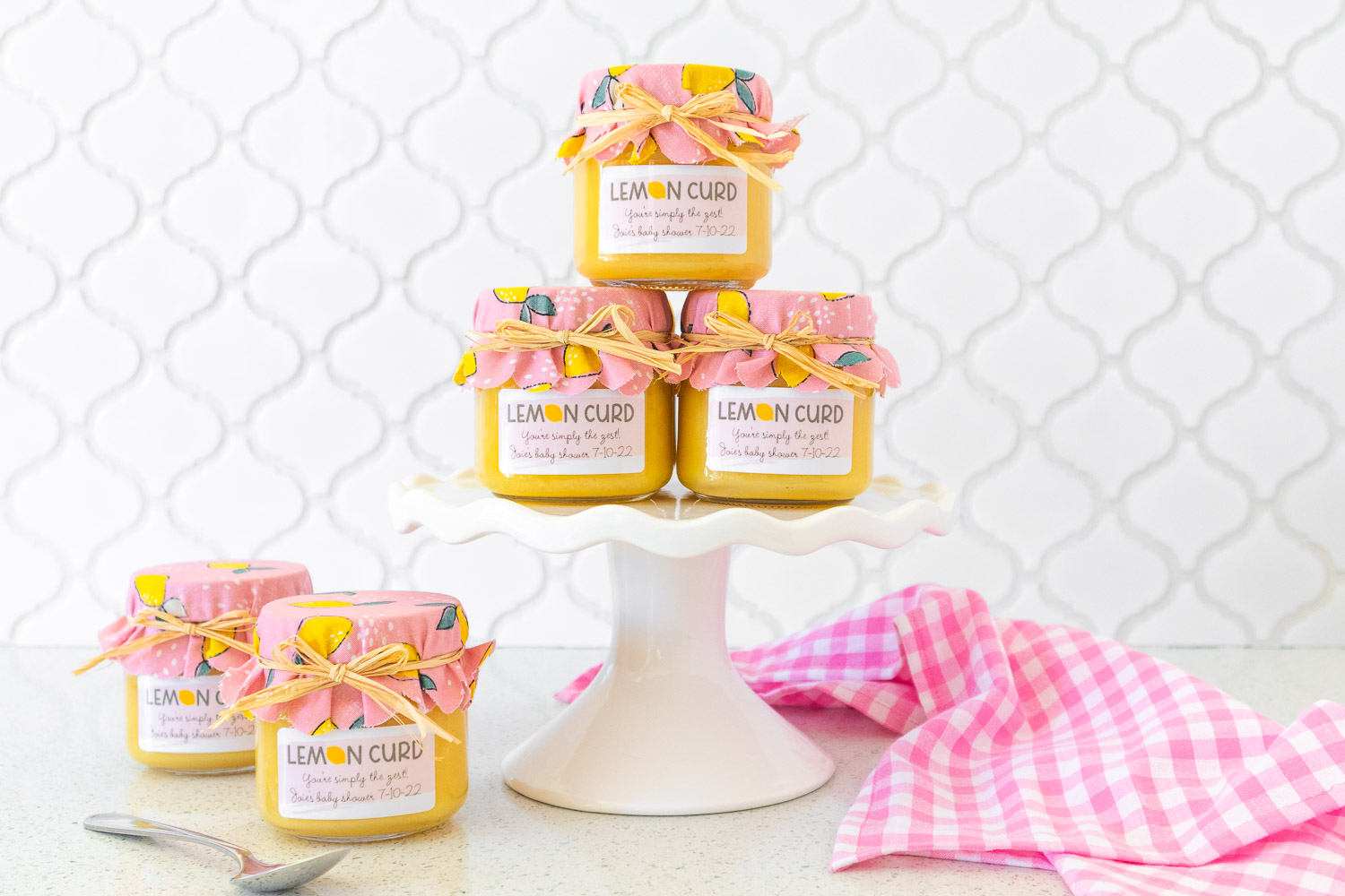 Lemon curd decorated with pink fabric jar covers and custom labels. A few are stacked on a white ceramic cake stand and two are off to the side. There's a bright pink gingham napkin in the right corner.