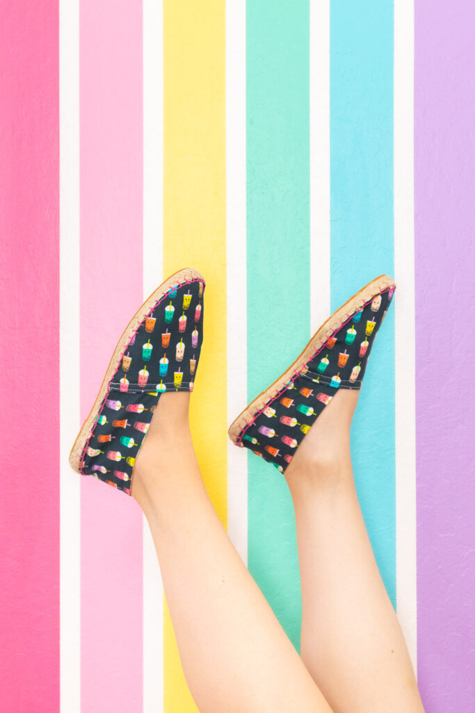 Close up of Blaire's feet are kicked up in the air. She is wearing espadrilles she made herself. They are navy with a colorful boba drink print. The sides are stitched with pink embroidery thread. The photo was taken against a rainbow wall backdrop. 