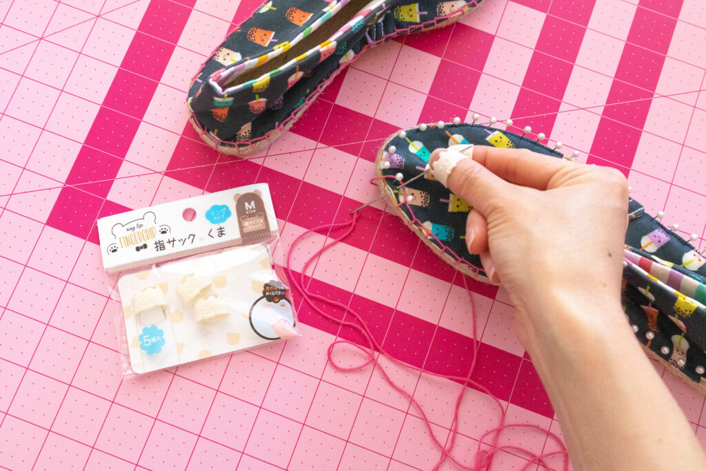 Sewing espadrille shoes with pink embroidery floss. Blaire's hand is in frame holding the needle with finger grips on her fingers. The shoes are navy with a rainbow bubble tea print.