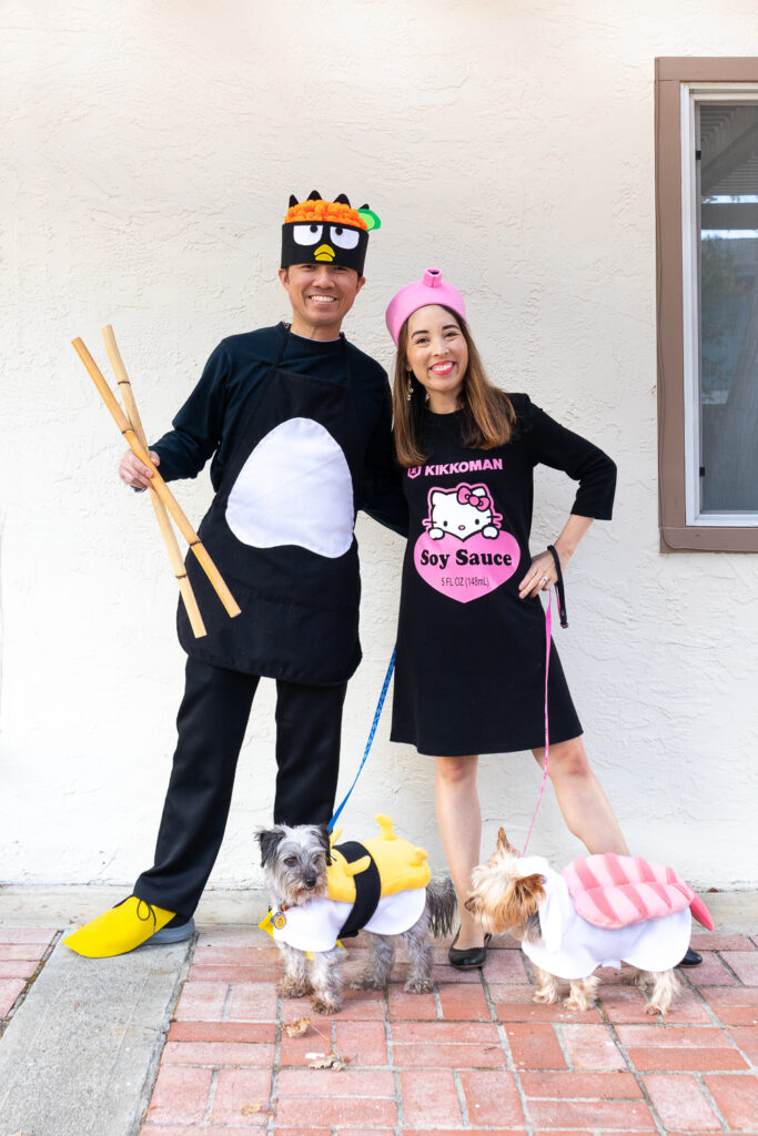 An Asian couple with their two small dogs dressed as Sanrio Sushi. The man is wearing a Badtz Maru chef costume, the woman is wearing a Hello Kitty x Kikkoman soy sauce costume. The gray, terrier mix dog is wearing a Gudetama tamago (egg) sushi costume. The tan Yorkie is wearing an ebi (shrimp) sushi costume. 