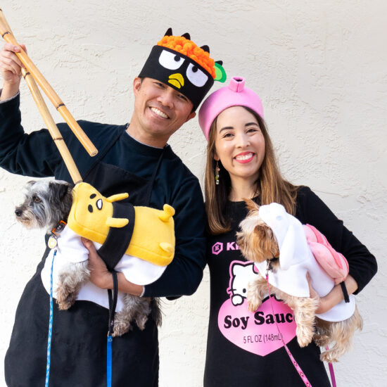An Asian couple with their two small dogs dressed as Sanrio Sushi. The man is wearing a Badtz Maru chef costume, the woman is wearing a Hello Kitty x Kikkoman soy sauce costume. The gray, terrier mix dog is wearing a Gudetama tamago (egg) sushi costume. The tan Yorkie is wearing an ebi (shrimp) sushi costume.