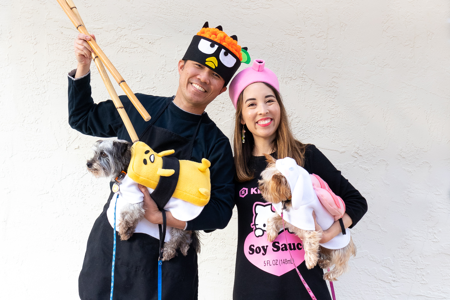 An Asian couple with their two small dogs dressed as Sanrio Sushi. The man is wearing a Badtz Maru chef costume, the woman is wearing a Hello Kitty x Kikkoman soy sauce costume. The gray, terrier mix dog is wearing a Gudetama tamago (egg) sushi costume. The tan Yorkie is wearing an ebi (shrimp) sushi costume.