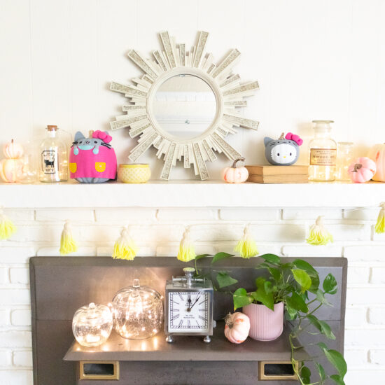 Off white wall and brick fireplace with mantel styled for Halloween. There are pastel, neon painted pumpkins, jars lit with fairy lights, Hello Kitty and Pusheen pumpkins on the mantel.