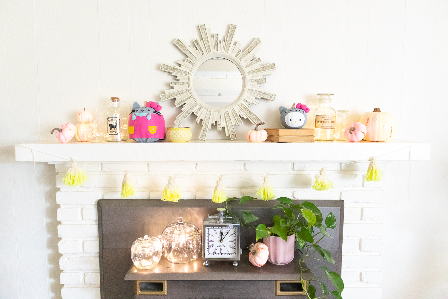 Off white wall and brick fireplace with mantel styled for Halloween. There are pastel, neon painted pumpkins, jars lit with fairy lights, Hello Kitty and Pusheen pumpkins on the mantel.