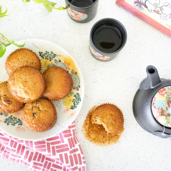 A flatlay photo of fuyu persimmon muffins on a colorful Japanese floral plate. Beneath the plate is a rose pink and white tea cloth. To the right of the plate is a muffin with a bite taken out of it and a Japanese tea pot and cup. There are also some green plants and a photo of bunnies poking into the frame.